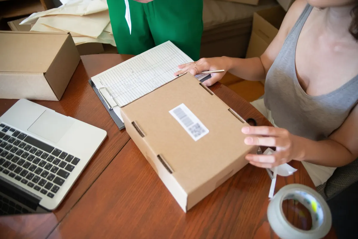 Two people at a table packing online orders, with a laptop, an order list, a labeled box and a roll of tape.