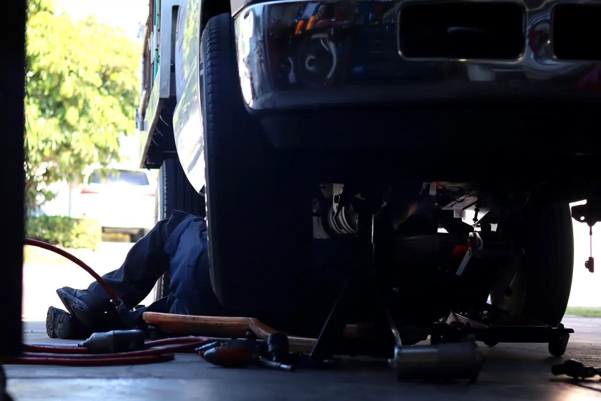 A technician on a creeper under a heavy-duty pickup, with an impact gun and air hose on the floor of an open shop bay.