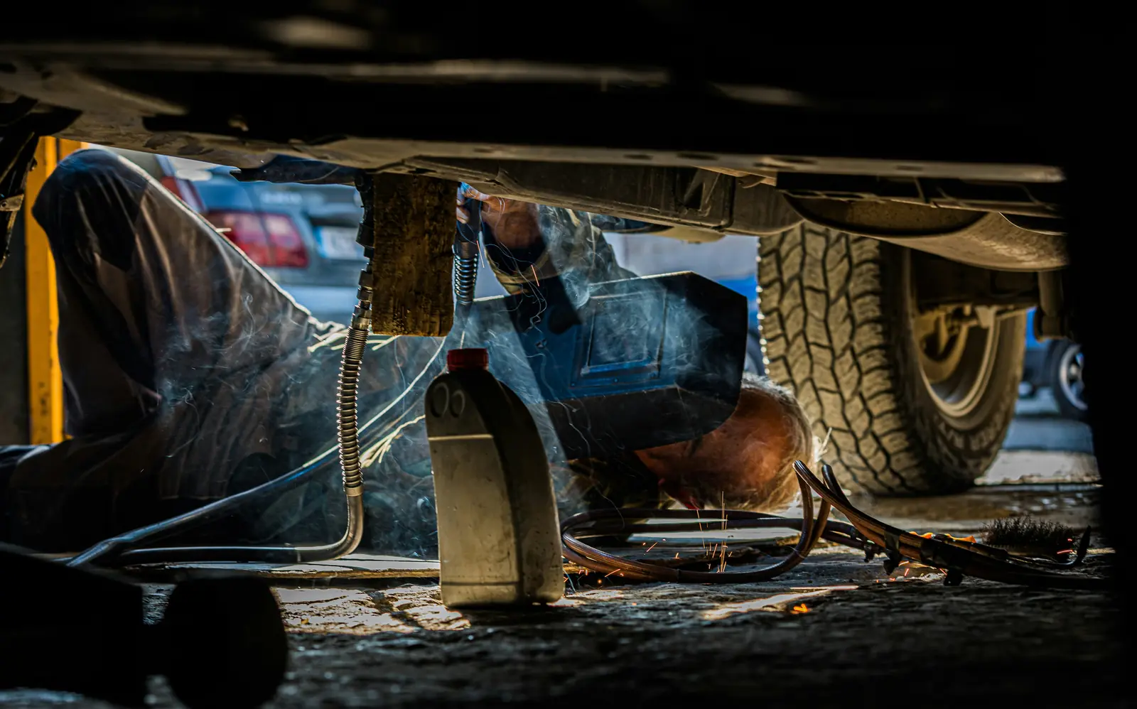 A mechanic lying on the shop floor under a pickup truck, welding, with a big all-terrain tire beside him.